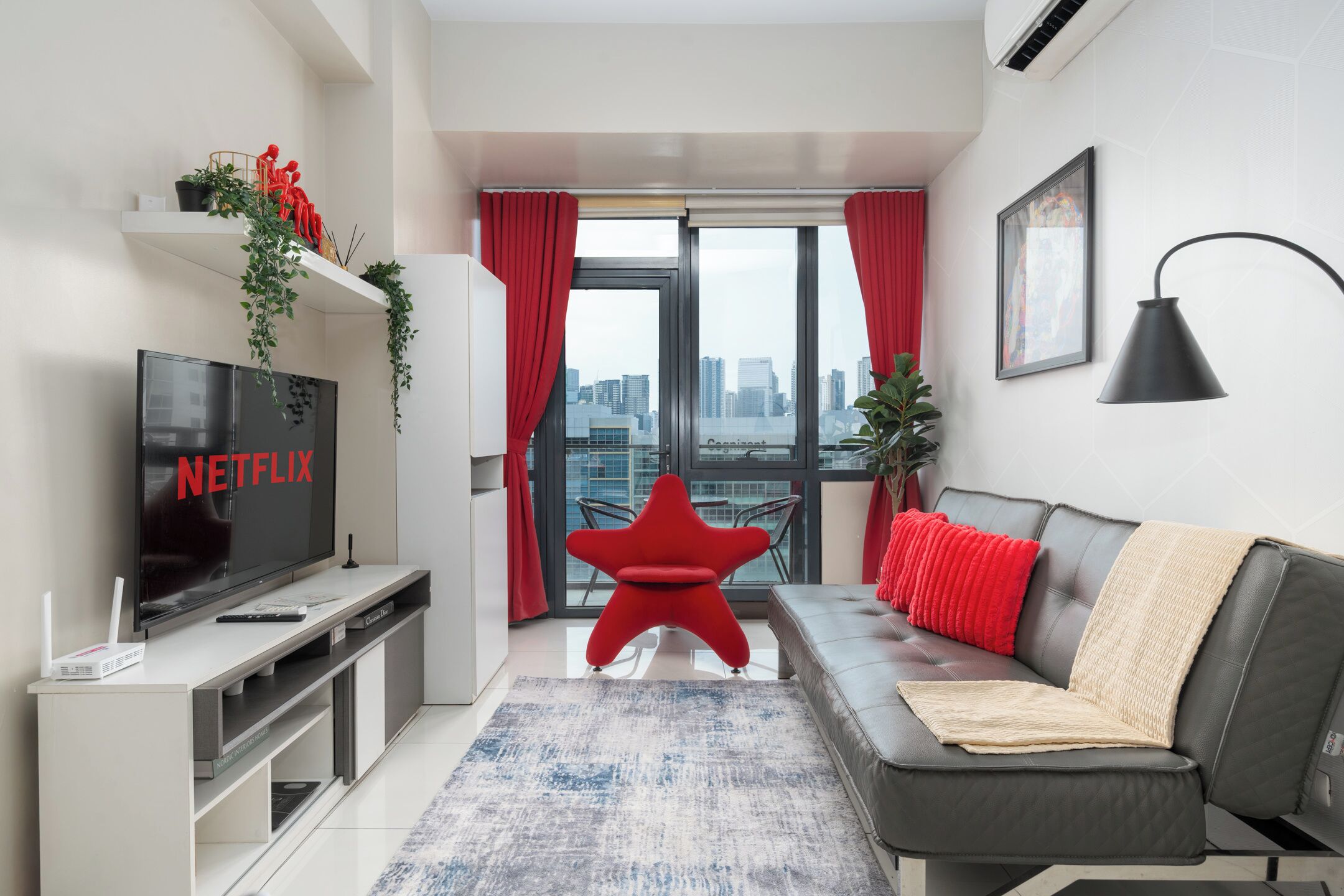 A modern living room with a flat-screen TV, a red chair, a grey sofa, and a city view through large windows.