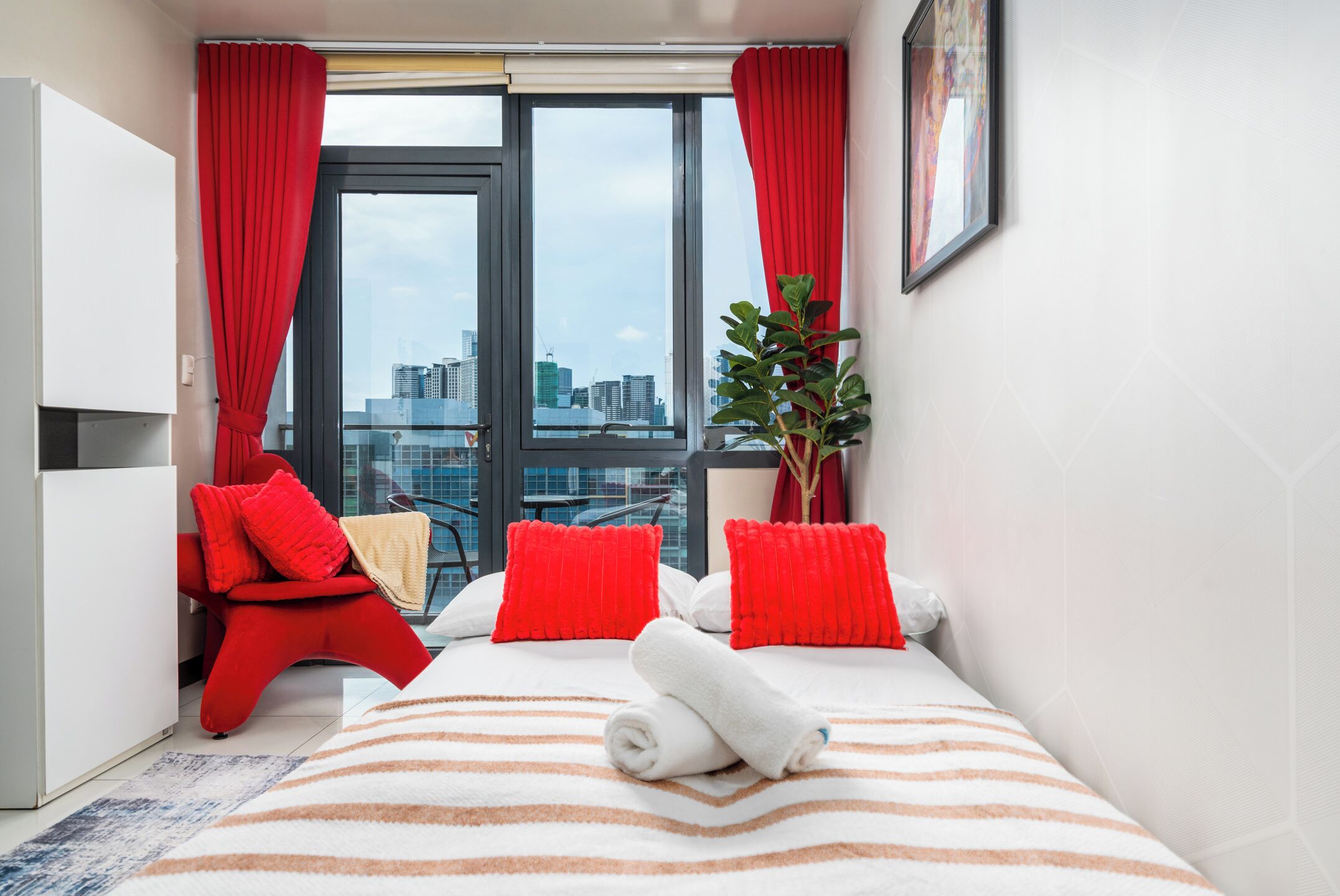 A hotel room with a bed, red pillows, a potted plant, and a city view.