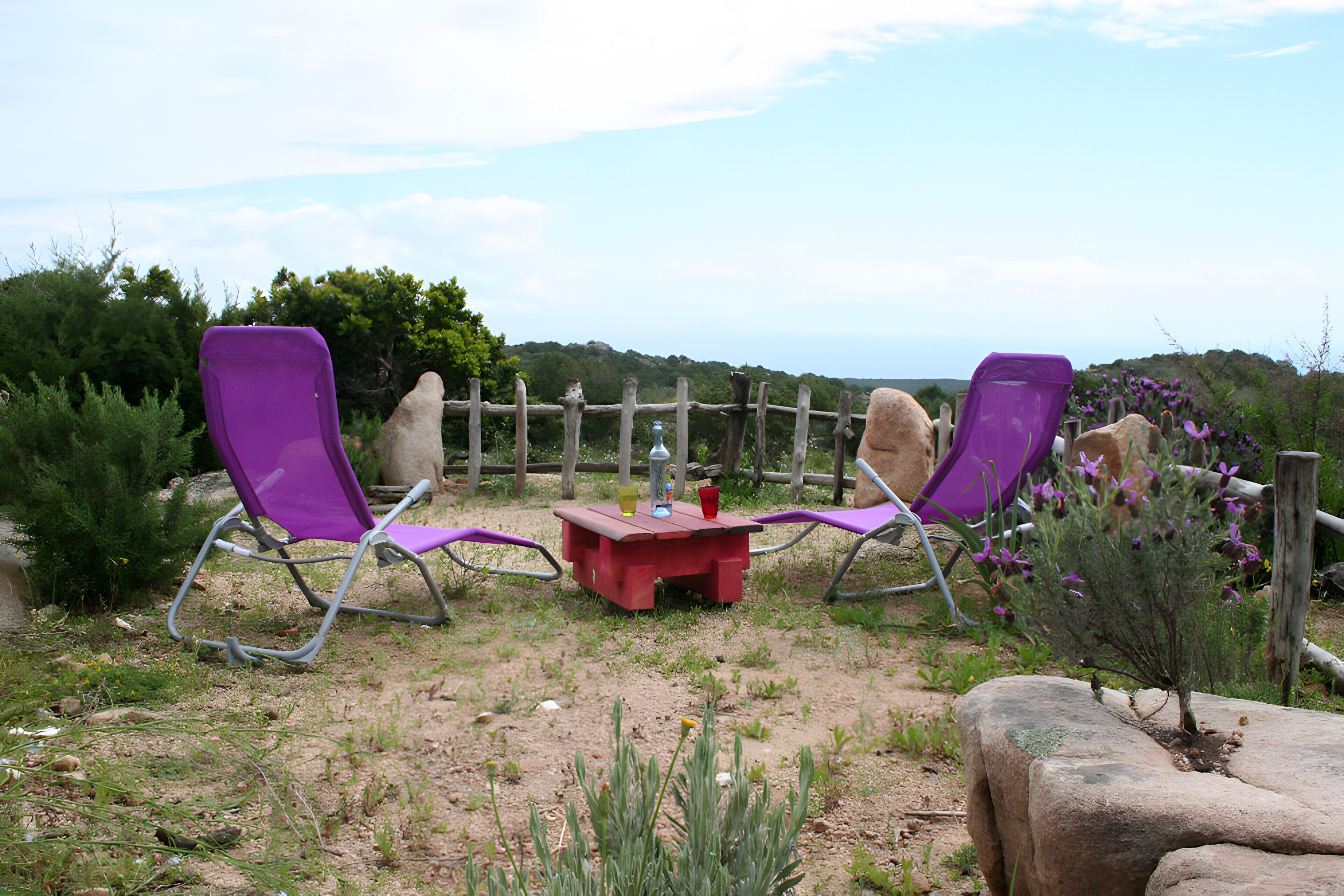 Twee paarse ligstoelen en een kleine rode tafel met drankjes, voor een achtergrond van een houten omheining en een natuurlandschap.