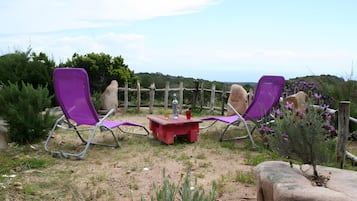 Twee paarse ligstoelen en een kleine rode tafel met drankjes, voor een achtergrond van een houten omheining en een natuurlandschap.