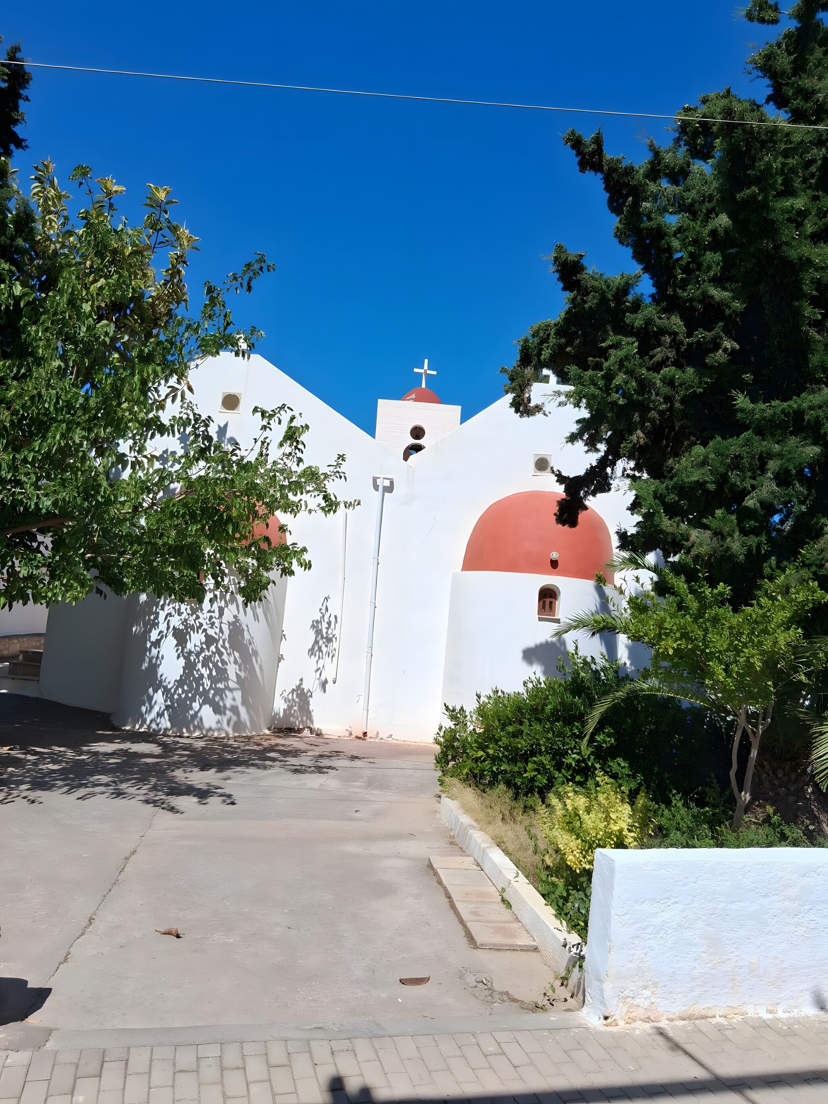 A white building with a red dome and a cross on top, surrounded by greenery.