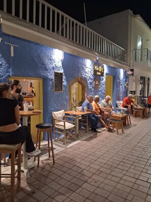 Un café al aire libre con mesas y sillas, un muro azul y una puerta amarilla.