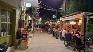 A paved street lined with shops and outdoor seating, with people dining and walking. - Apartment 'Porto Beach' with Sea View, Wi-Fi and Air Conditioning (Makry Gialos)