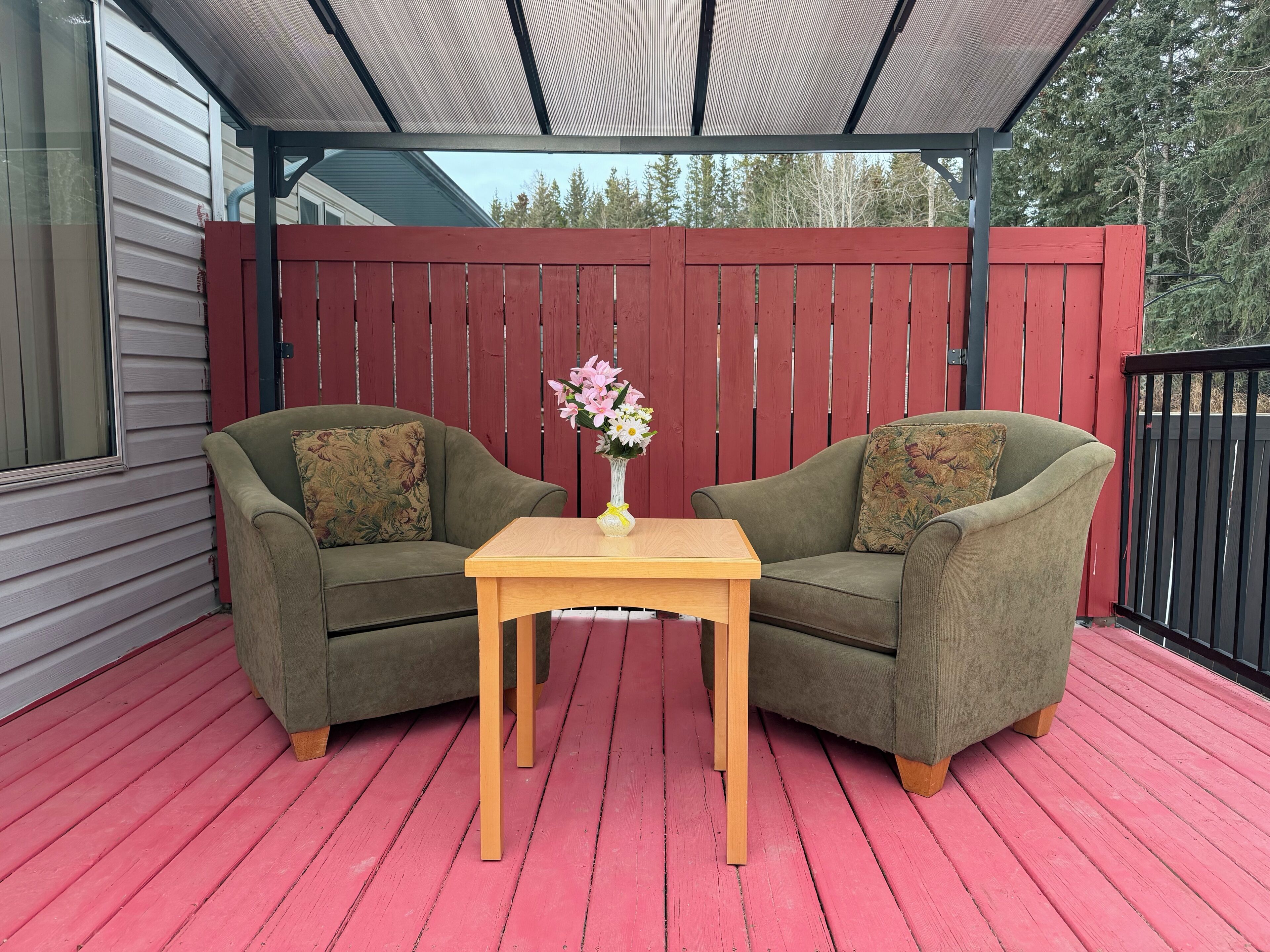 A covered outdoor seating area with two green armchairs and a wooden table, a red fence, and a house in the background.