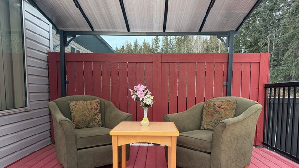 Un área de estar al aire libre con techo, con dos sillones verdes y una mesa de madera, una cerca roja y una casa al fondo.
