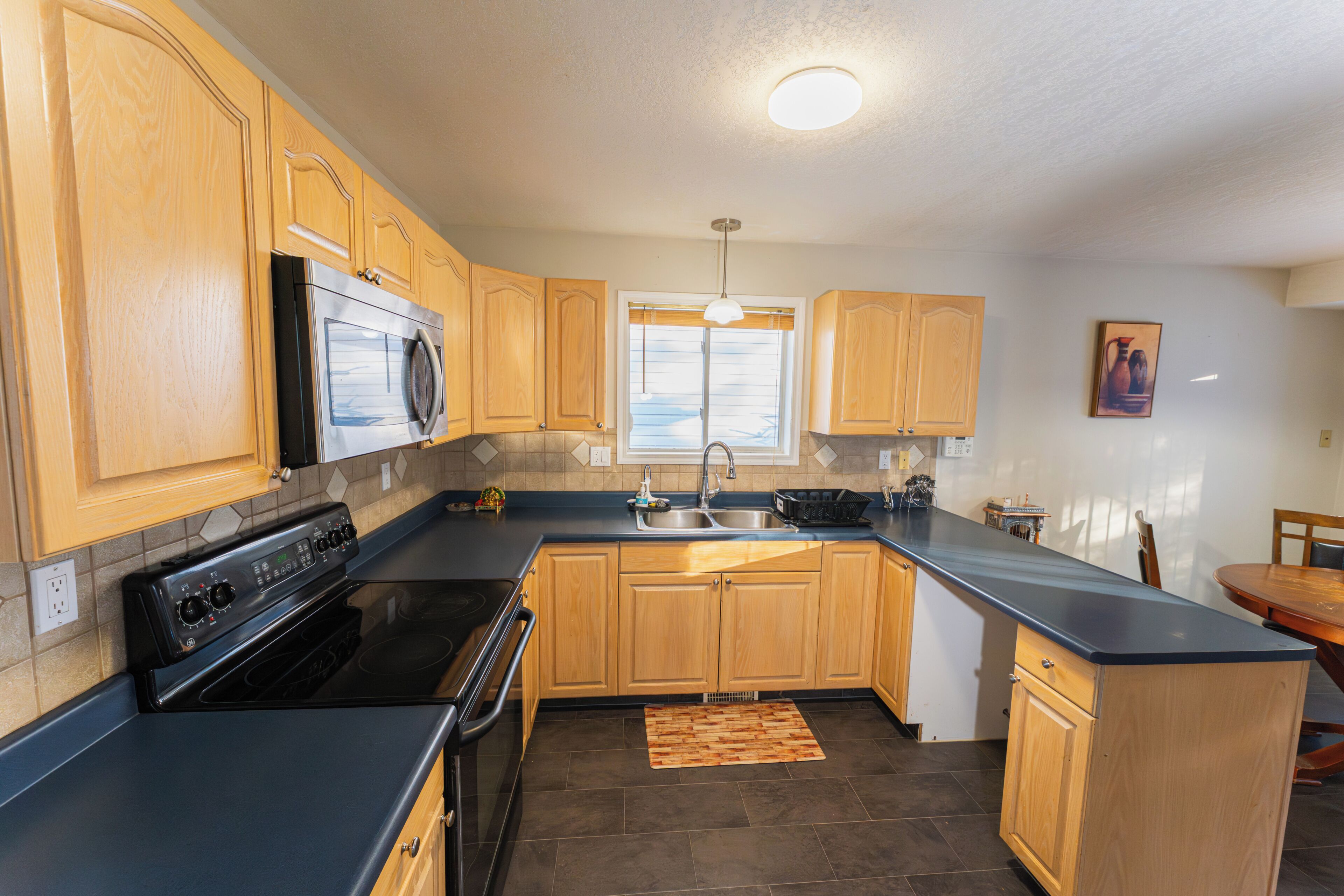 A kitchen with wooden cabinets, a black countertop, a stainless steel oven, and a window with blinds.