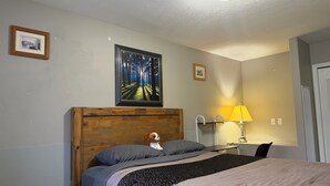 Un dormitorio con cabecera de madera, cama con ropa de cama gris, mesita de noche con lámpara, y un cuadro enmarcado en la pared.