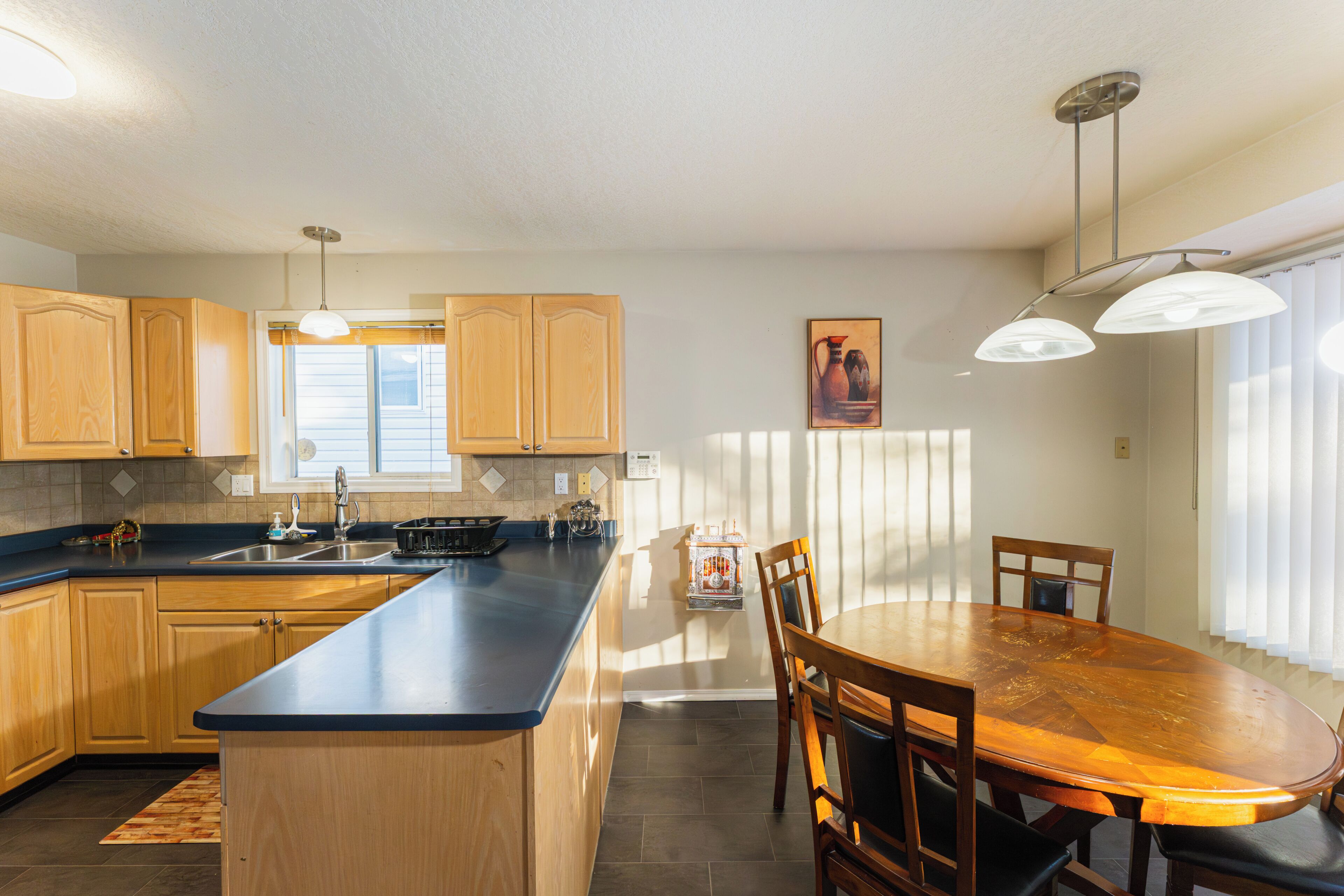 A kitchen with wooden cabinets, a central island, a dining table with chairs, pendant lights, and a window with blinds.