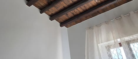 A room with a wooden ceiling, a flat-screen TV mounted on the wall, and a window with sheer curtains.