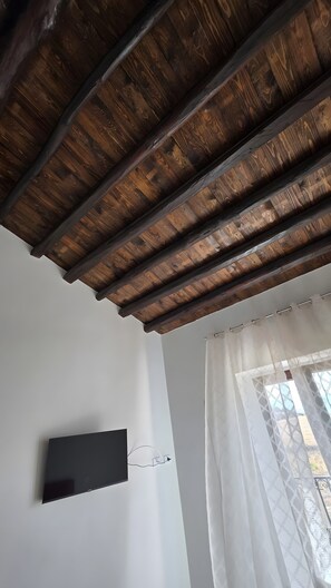 A room with a wooden ceiling, a flat-screen TV mounted on the wall, and a window with sheer curtains. - Solerata Room – Sikania Agrirelais, Mountain View (Caccamo)