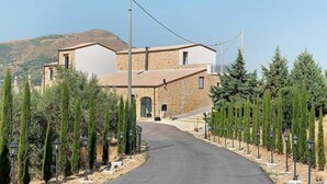 Un edificio in pietra su due piani con un ingresso centrale, affiancato da file di alberi di conifere, che conduce a un vialetto lastricato.