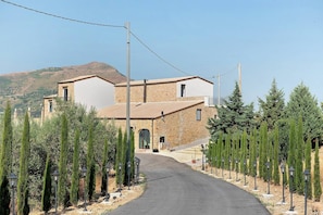 A two-story stone building with a central entrance, flanked by rows of coniferous trees, leading to a paved driveway. - Solerata Room – Sikania Agrirelais, Mountain View (Caccamo)