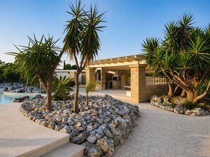 A poolside area with a stone structure, palm trees, and a clear sky. - SeaShell – Salento Retreat: Private Pool, Wi-Fi & A/C, 1 km from the Sea (Corsano)