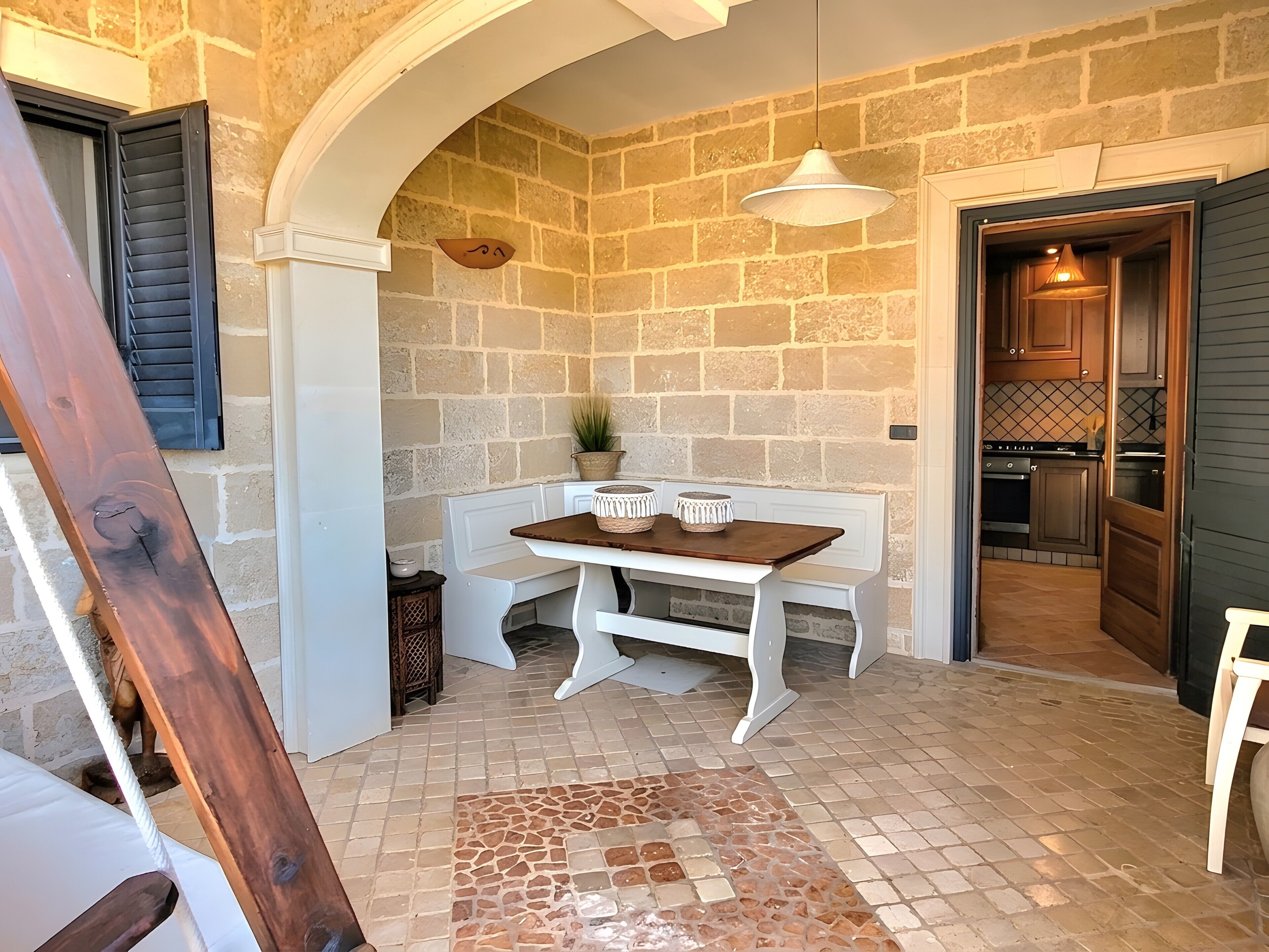 A stone-walled room with a wooden bench, a hanging light fixture, and a view into the kitchen.
