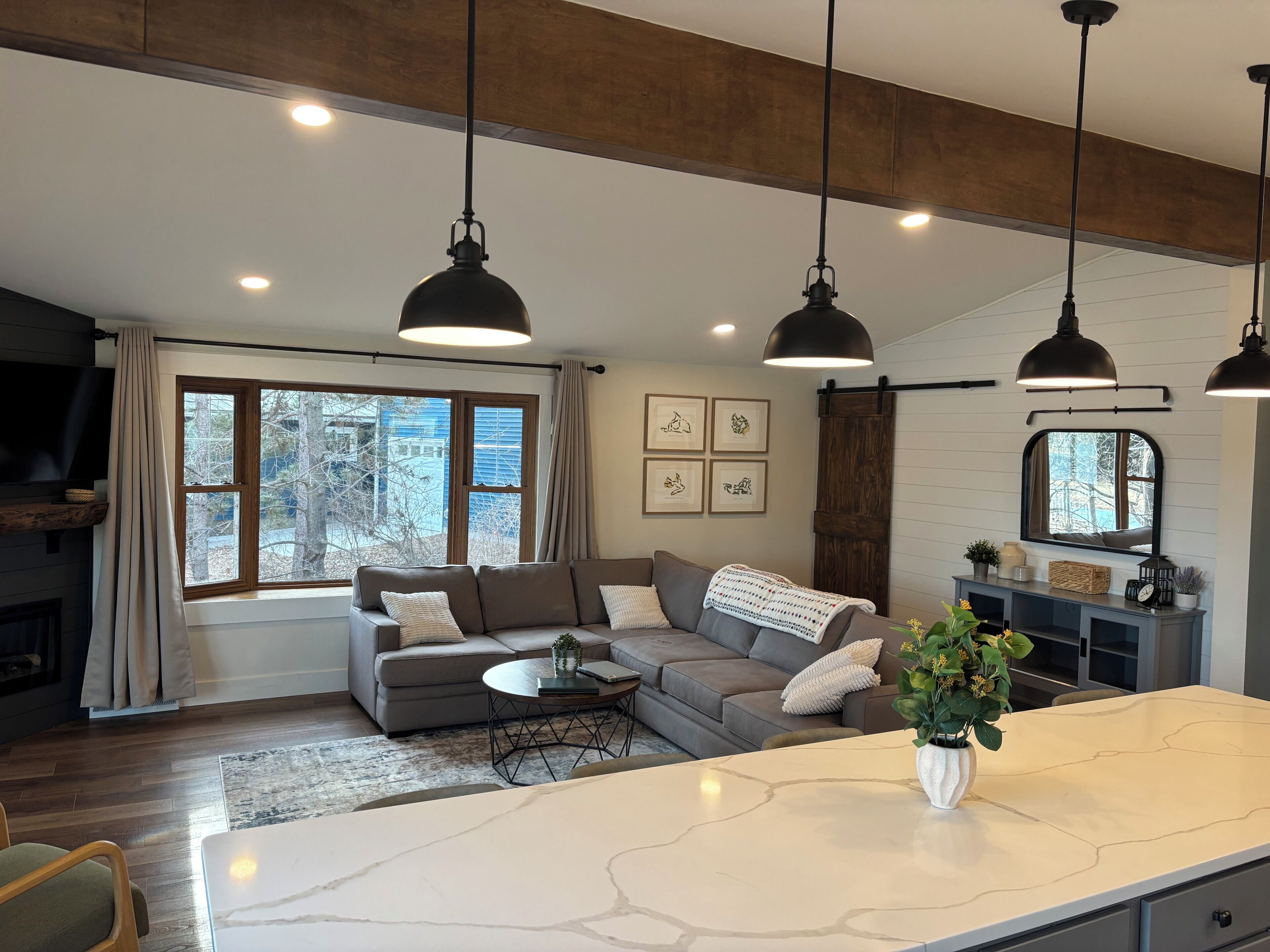 A modern living room with a sofa, a coffee table, pendant lights, and a large window with a view of trees.