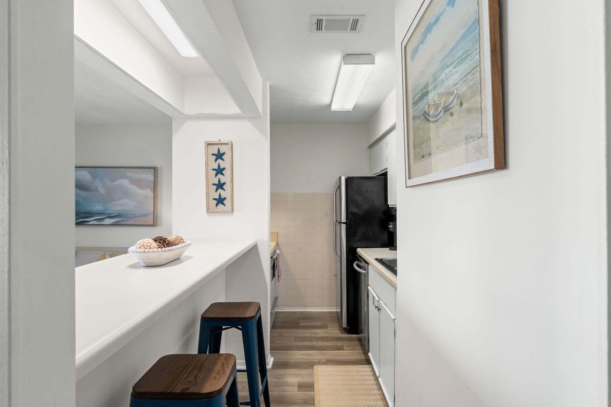 Une cuisine étroite avec un plan de travail blanc, un réfrigérateur noir et un tabouret en bois.
