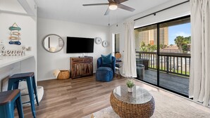 Ein modernes Wohnzimmer mit Fernseher, Holzmöbeln, Deckenventilator und einem Balkon mit Blick auf Gebäude und Palmen.