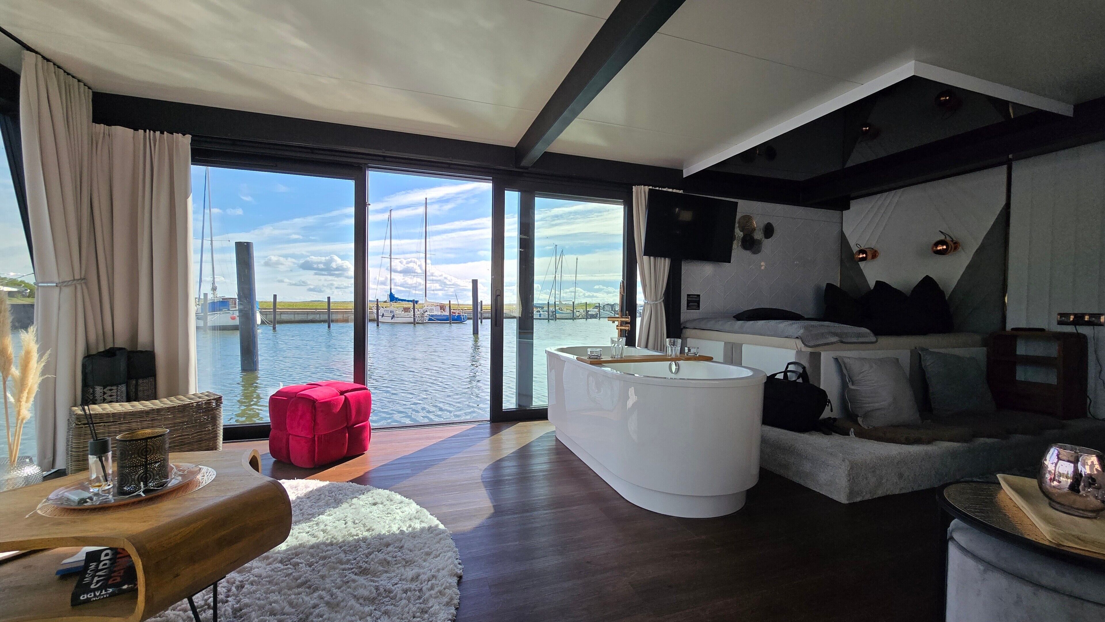 A modern interior with a large window offering a marina view, a white bathtub, a sofa, and a flat-screen TV mounted on the wall.