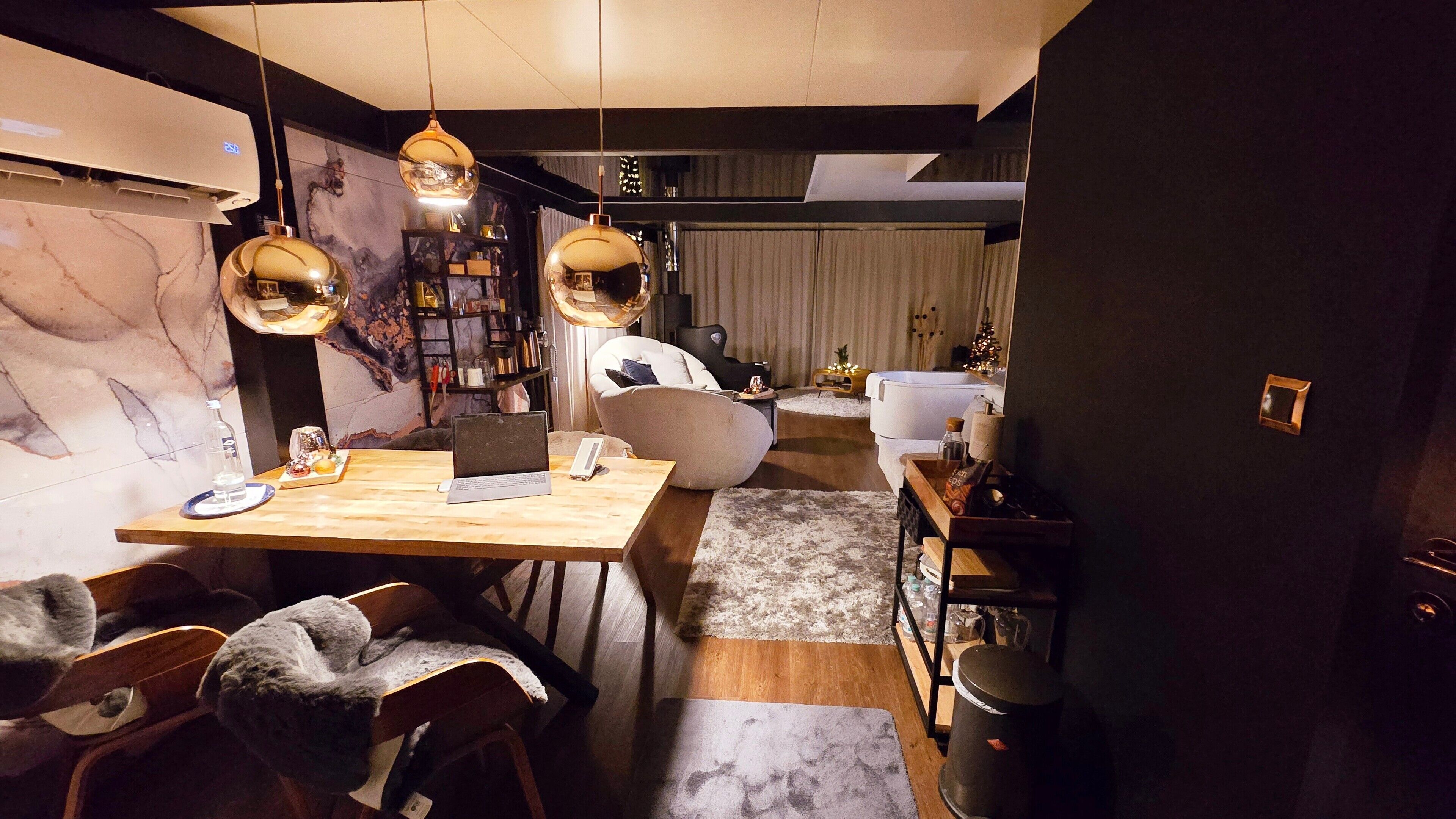 Et moderne spiseområde med et træbord, stole og en bærbar computer. Der er en reol med bøger og dekorative genstande, samt en hvid sofa i baggrunden.