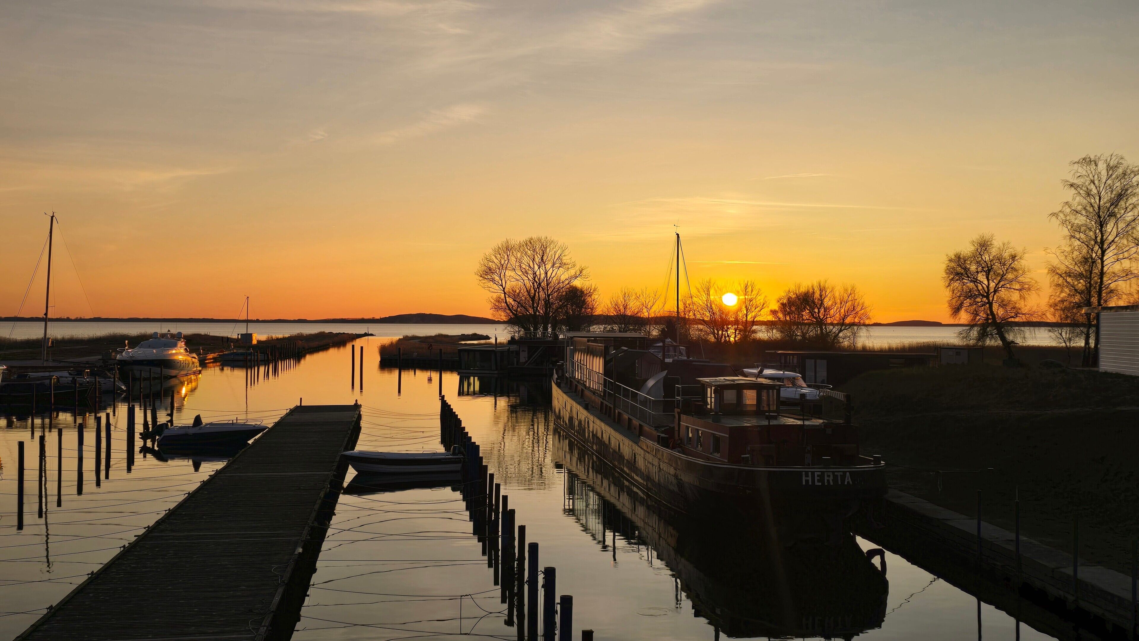 A calm waterway with boats docked at a pier, a clear sky, and a sunset.