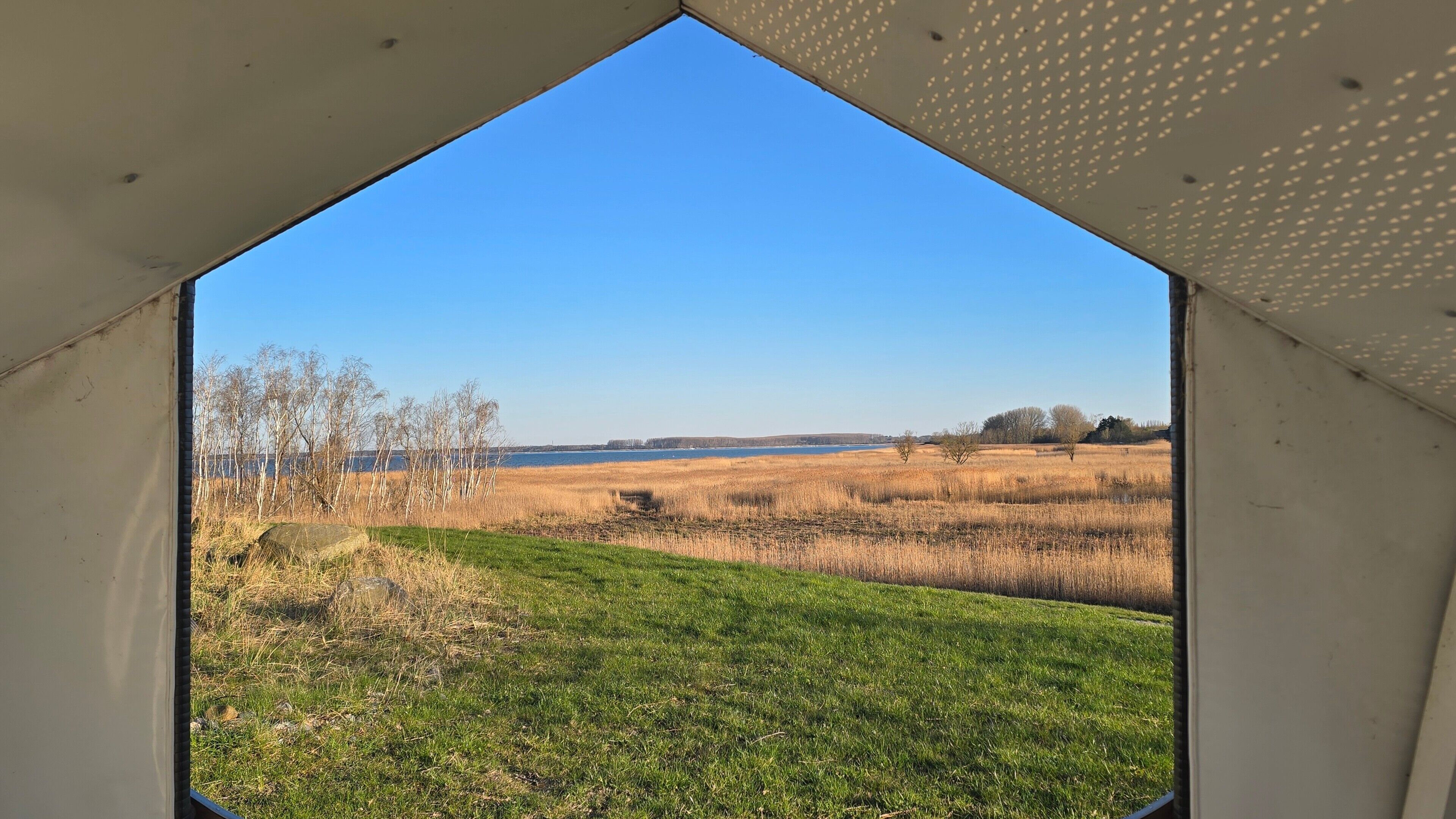 Blick durch ein dreieckiges Fenster auf ein Wiesenfeld mit einem Gewässer in der Ferne.
