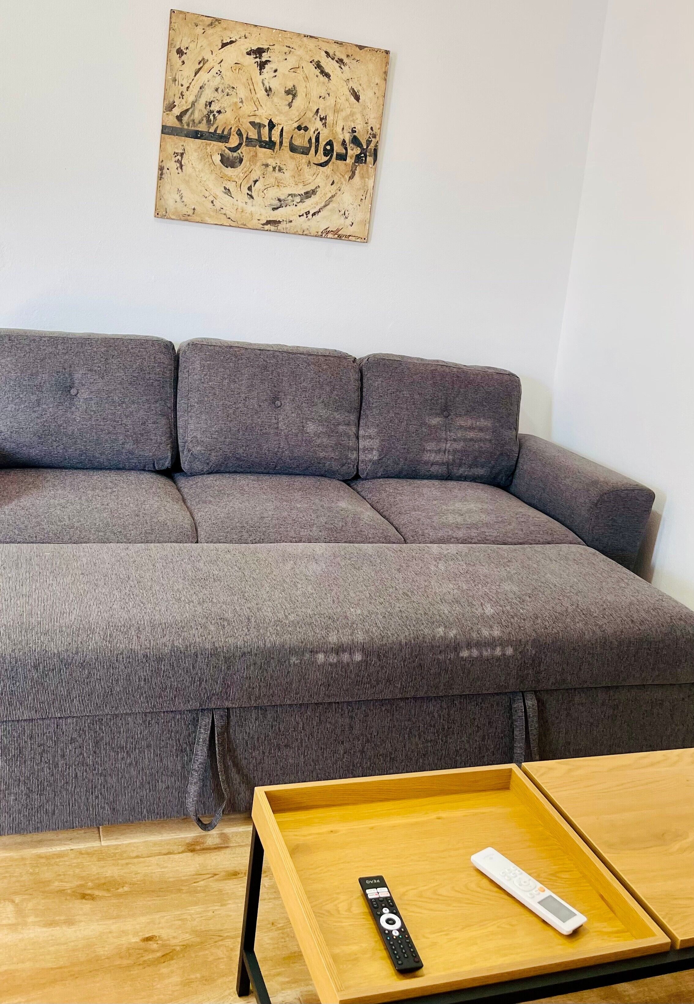 Un sofá gris con un plaid estampado, una mesa de centro de madera con un control remoto y una obra de arte colgada en la pared.