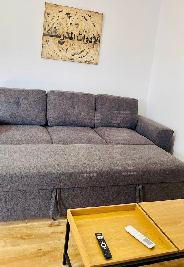 Un canapé gris avec un plaid à motifs, une table basse en bois avec une télécommande, et une œuvre d’art accrochée au mur.