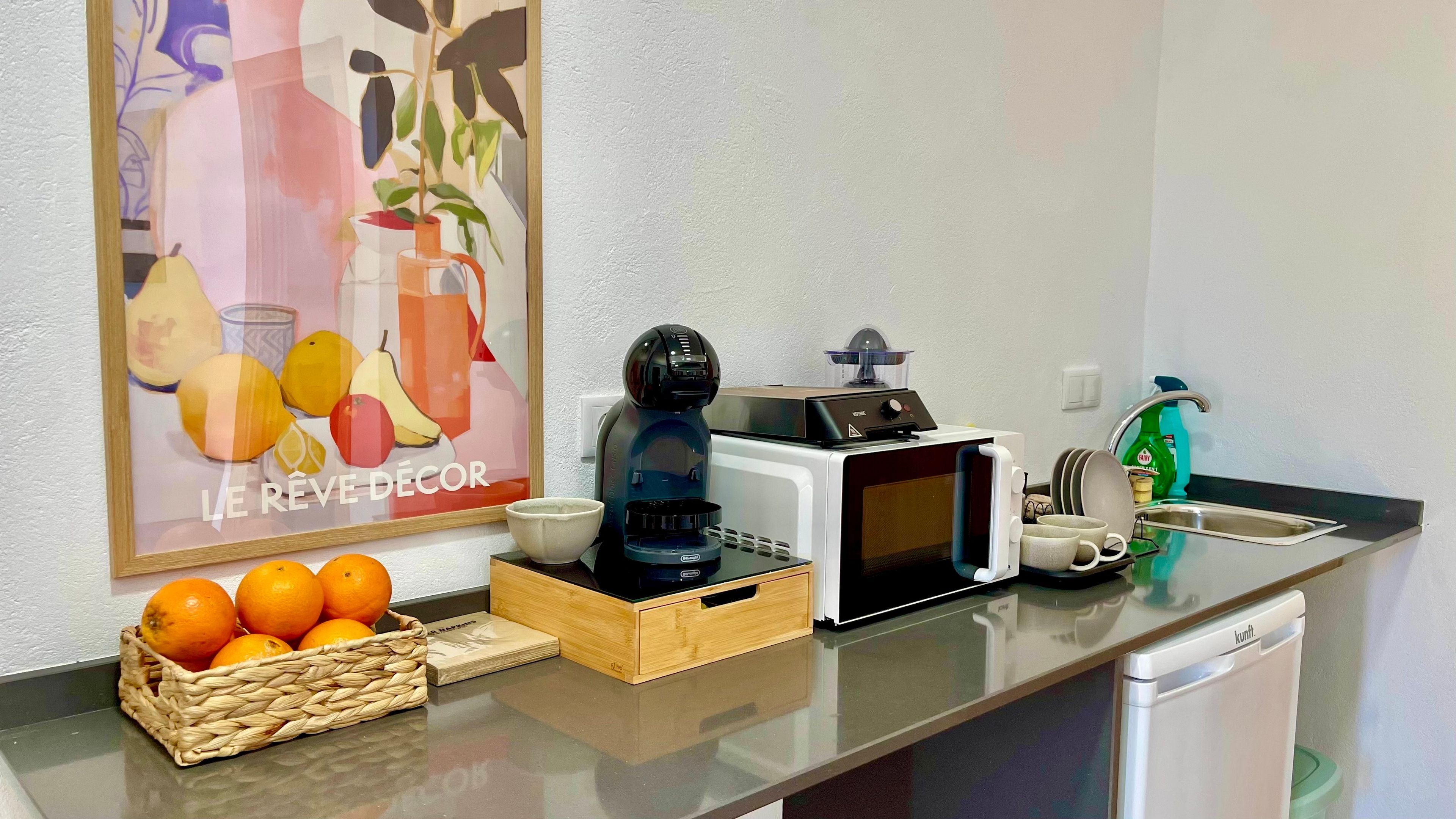A kitchen counter with a coffee maker, microwave, and dish rack. There is a basket of oranges and a framed picture on the wall.