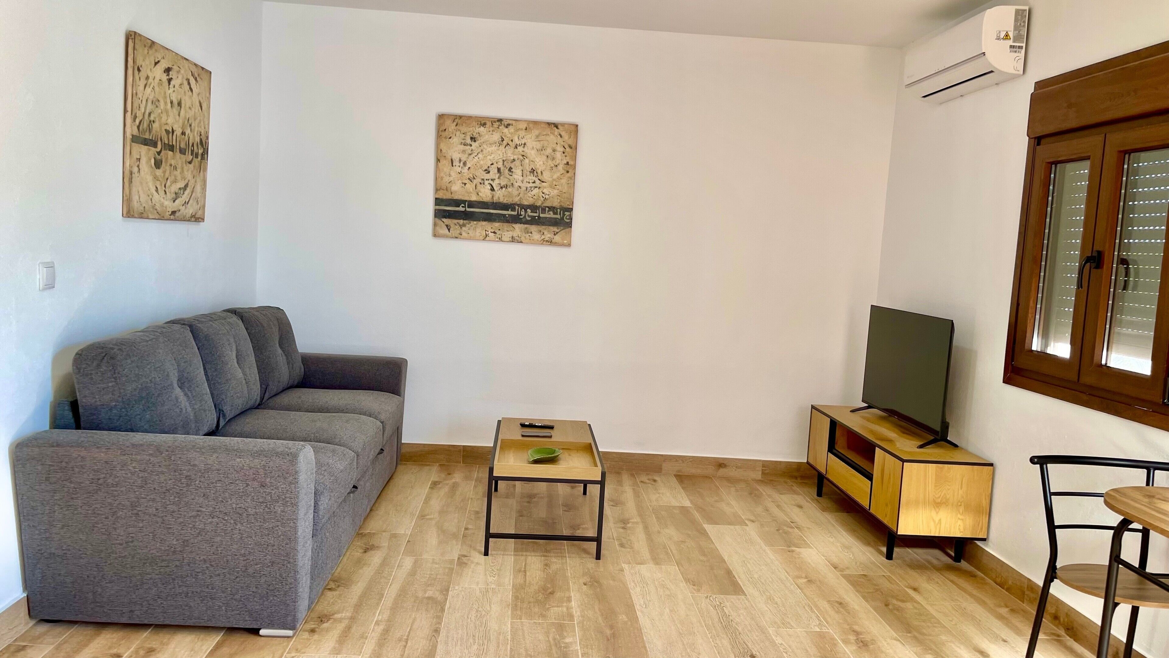 Ein Wohnzimmer mit einem grauen Sofa, einem kleinen Holztisch, einem Fernseher auf einem Holzständer und einem Fenster mit Jalousien.