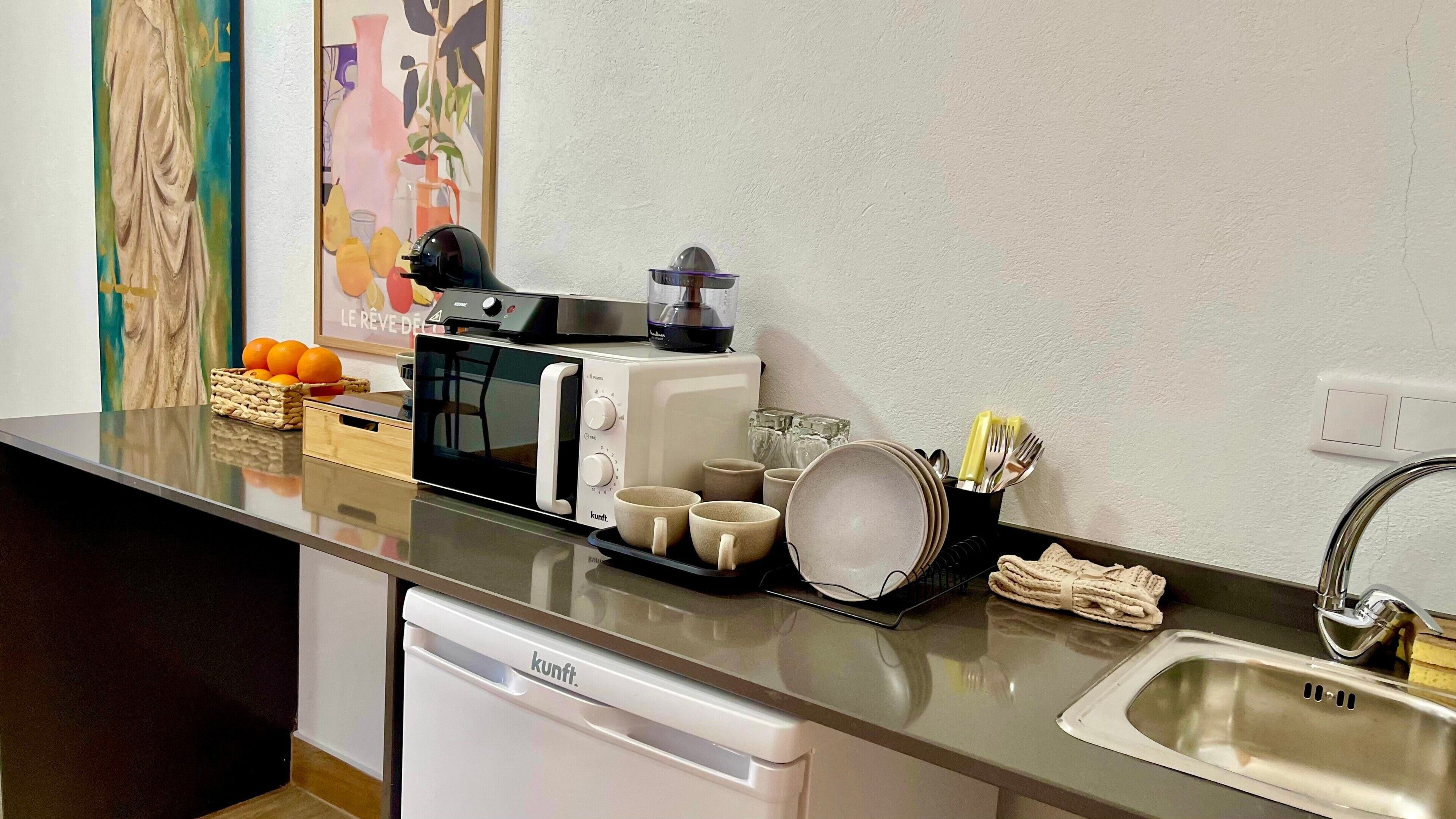 Una kitchenette con microondas, tostadora y hervidor. Hay tazas, una cesta de naranjas y un lavabo con grifo.