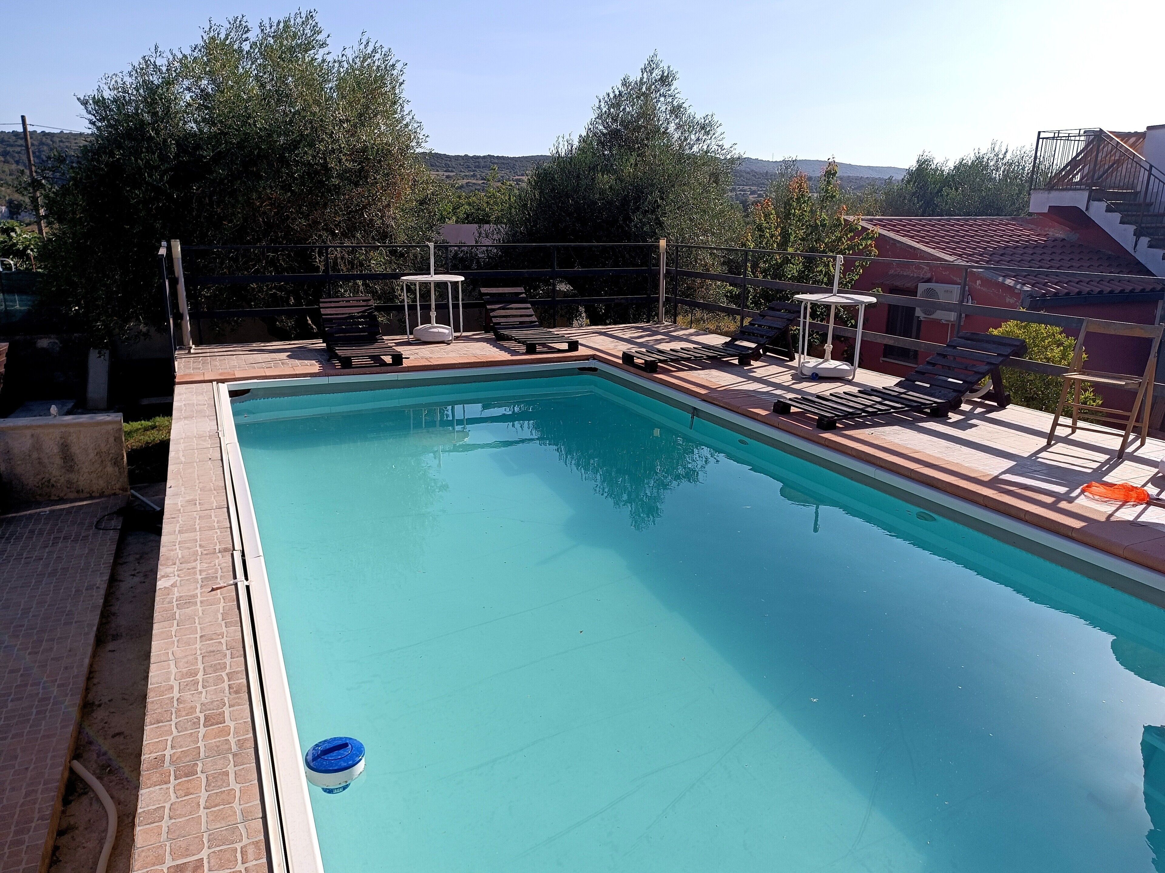 A rectangular swimming pool with a tiled edge, surrounded by a brick deck. There are lounge chairs and a sunshade structure.