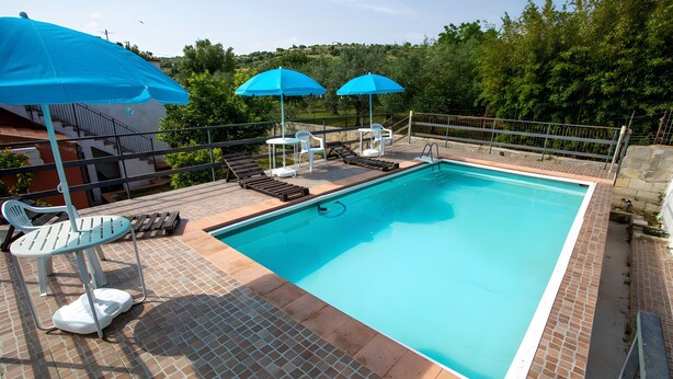 A rectangular swimming pool with a tiled deck, surrounded by a metal railing. There are lounge chairs and umbrellas, and a small table with chairs. - Villa 'La Panchina' with private pool, Wi-Fi, and air conditioning (Noto)
