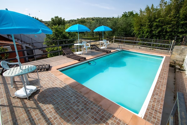 Ein rechteckiger Pool mit Fliesen umgeben, mit einem Metallgeländer. Liegestühle und Sonnenschirme sind vorhanden, sowie ein kleiner Tisch mit Stühlen.