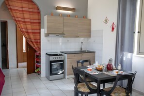 A compact kitchen with a stove, oven, and sink, a dining table with chairs, and a window with curtains.