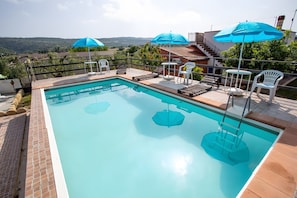 Ein rechteckiger Pool mit glasklarer, blauer Wasseroberfläche, umgeben von einer gefliesten Liegefläche mit Liegestühlen und Sonnenschirmen.