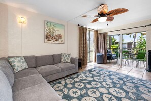 A living room with a grey sectional sofa, a patterned rug, a ceiling fan, and a painting of seashells. - 🏖️Top-Floor Kona Condo with A/C & Private Lanai (Kailua Kona)