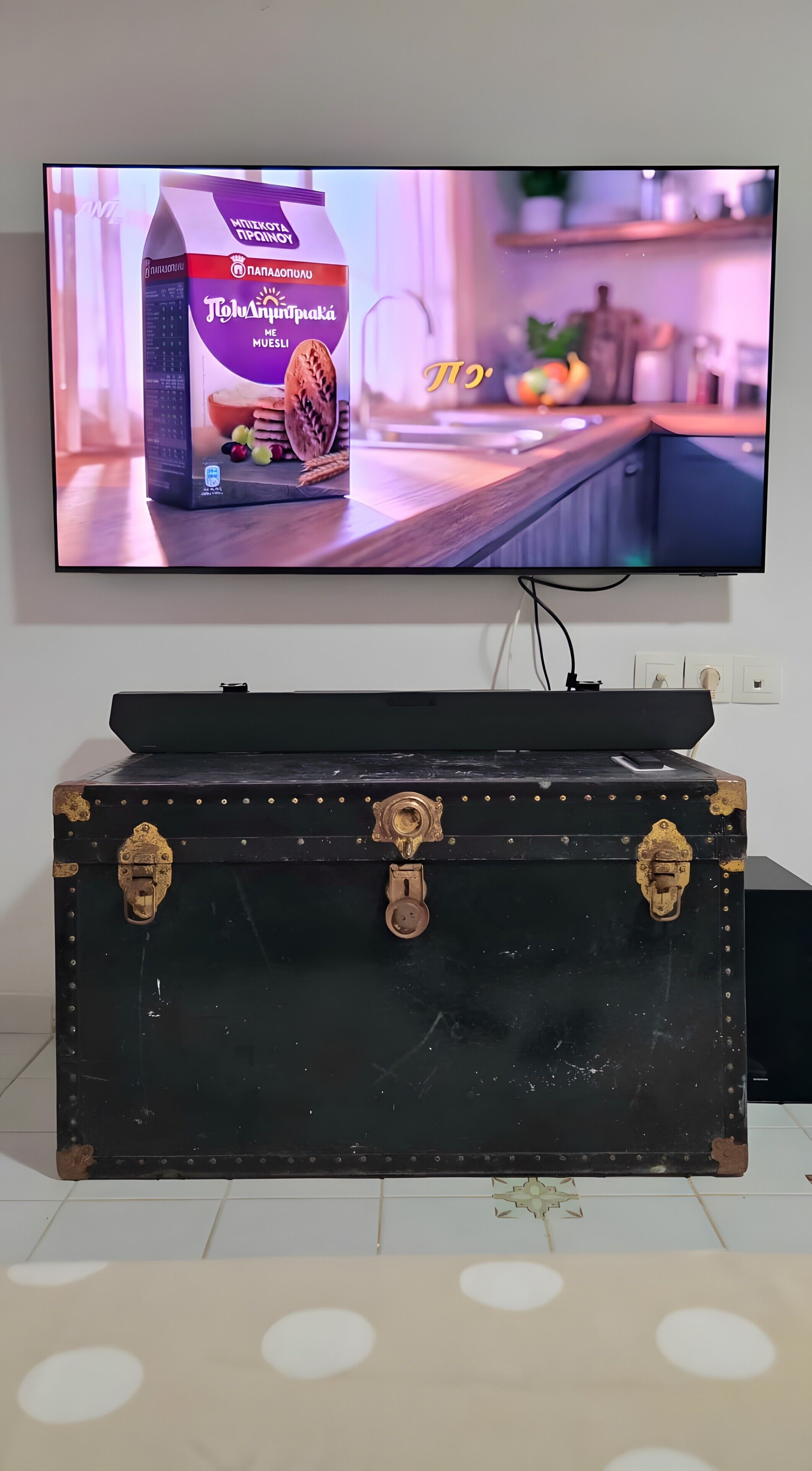 Un televisore che mostra una pubblicità di un prodotto, un baule d'epoca in primo piano e un angolo cottura sullo sfondo.
