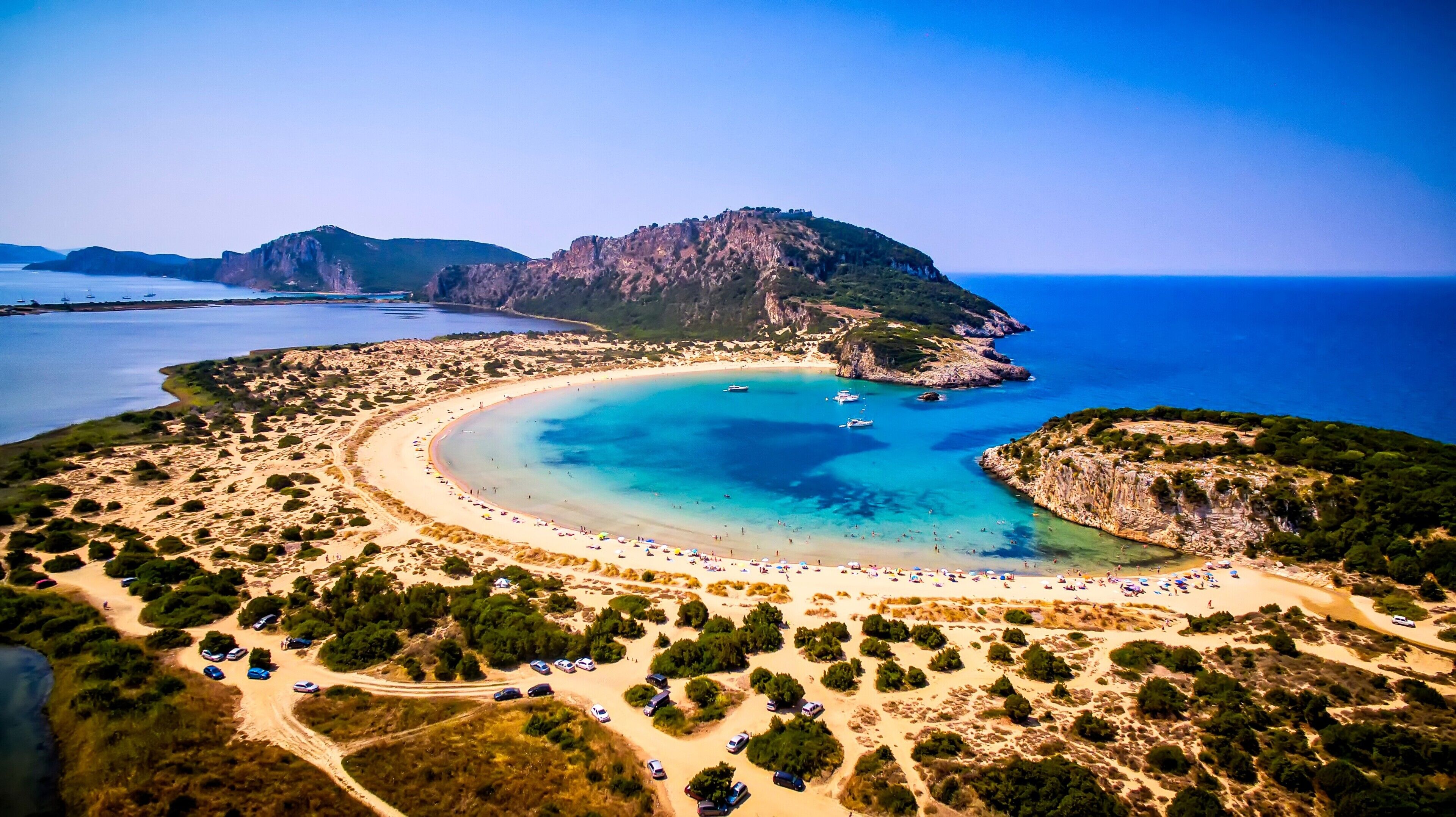 An aerial view of a coastal area with a beach, boats, and a mountainous backdrop.