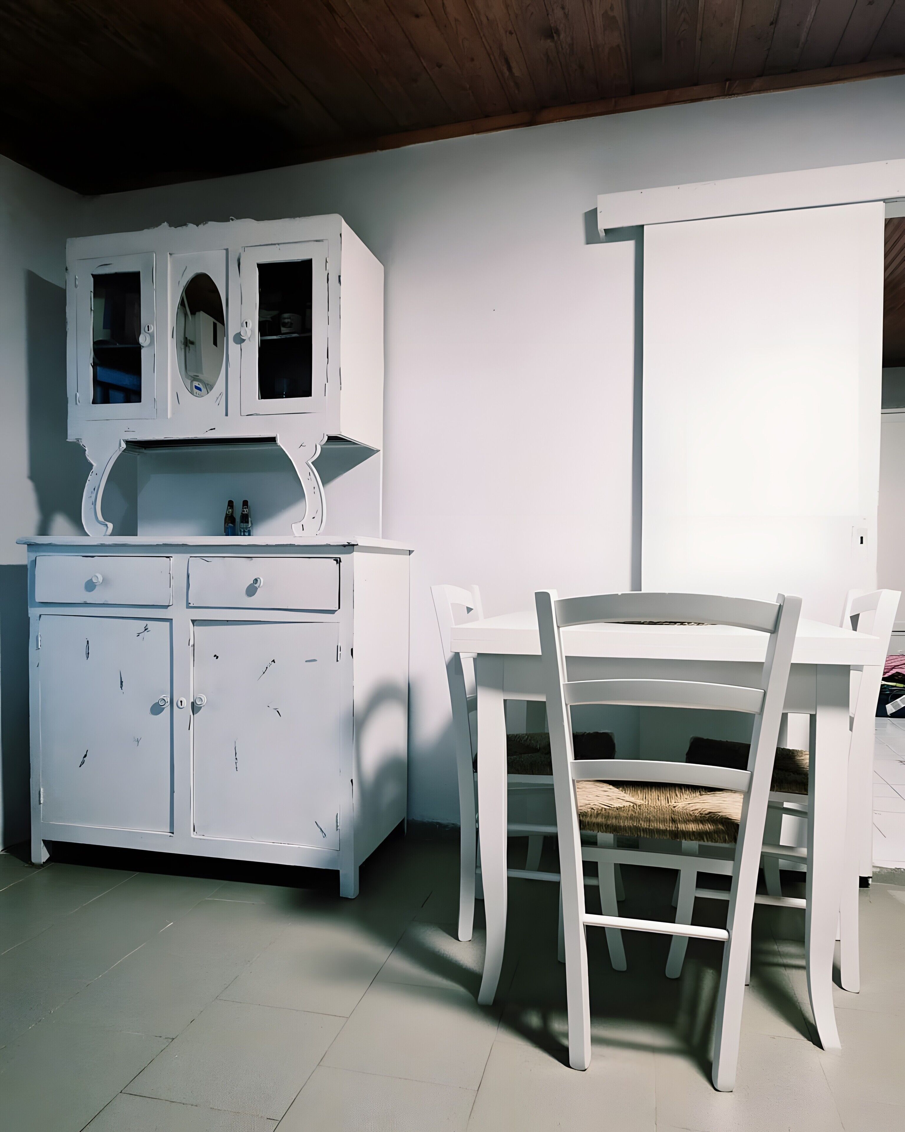 A white dining area with a table and chairs, a white cabinet with two doors, and a wooden ceiling.