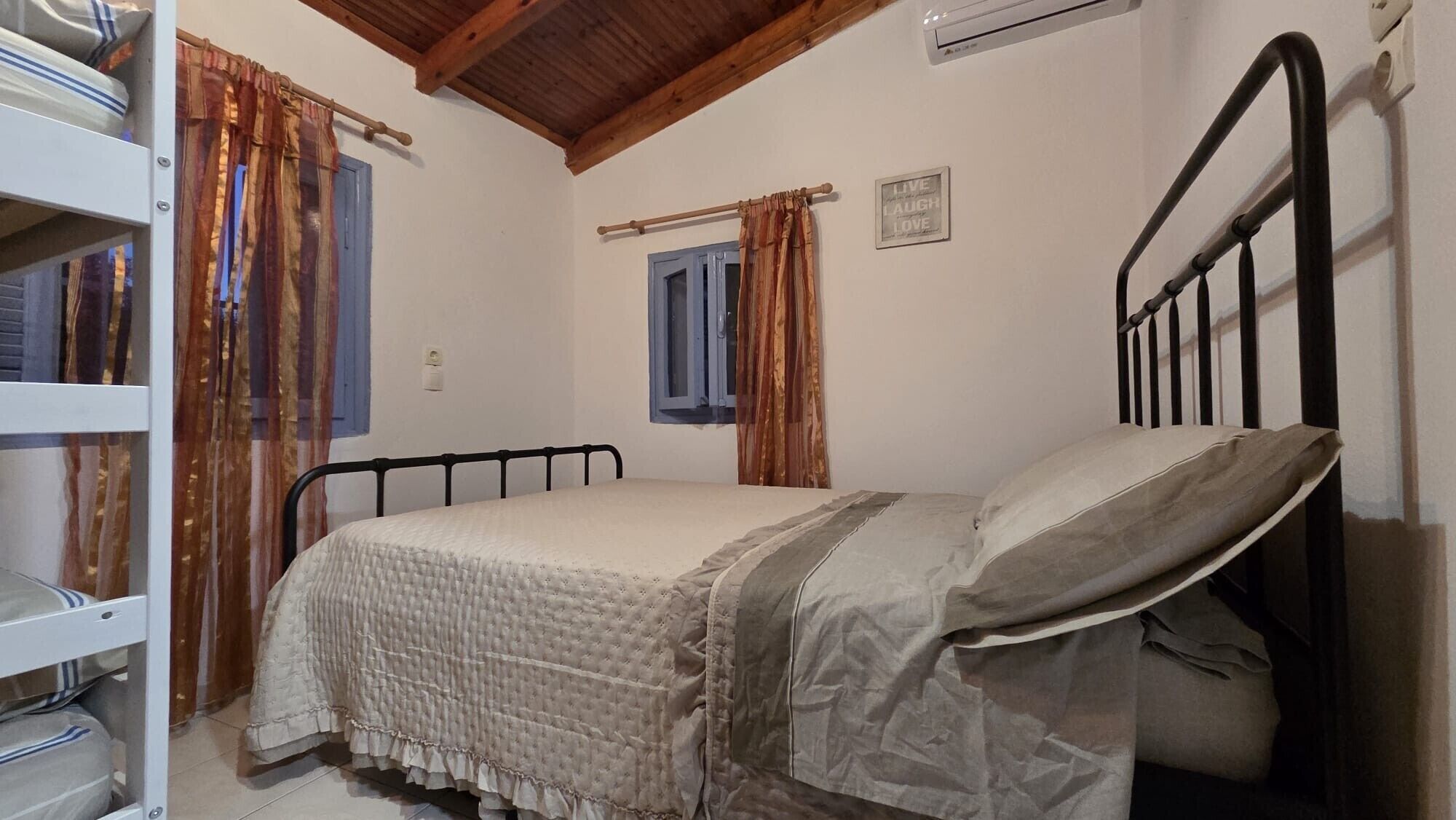 Una cama individual con cabecera, una ventana con cortinas, un aire acondicionado instalado en la pared y un letrero que dice 'LIVE LAUGH LOVE'.