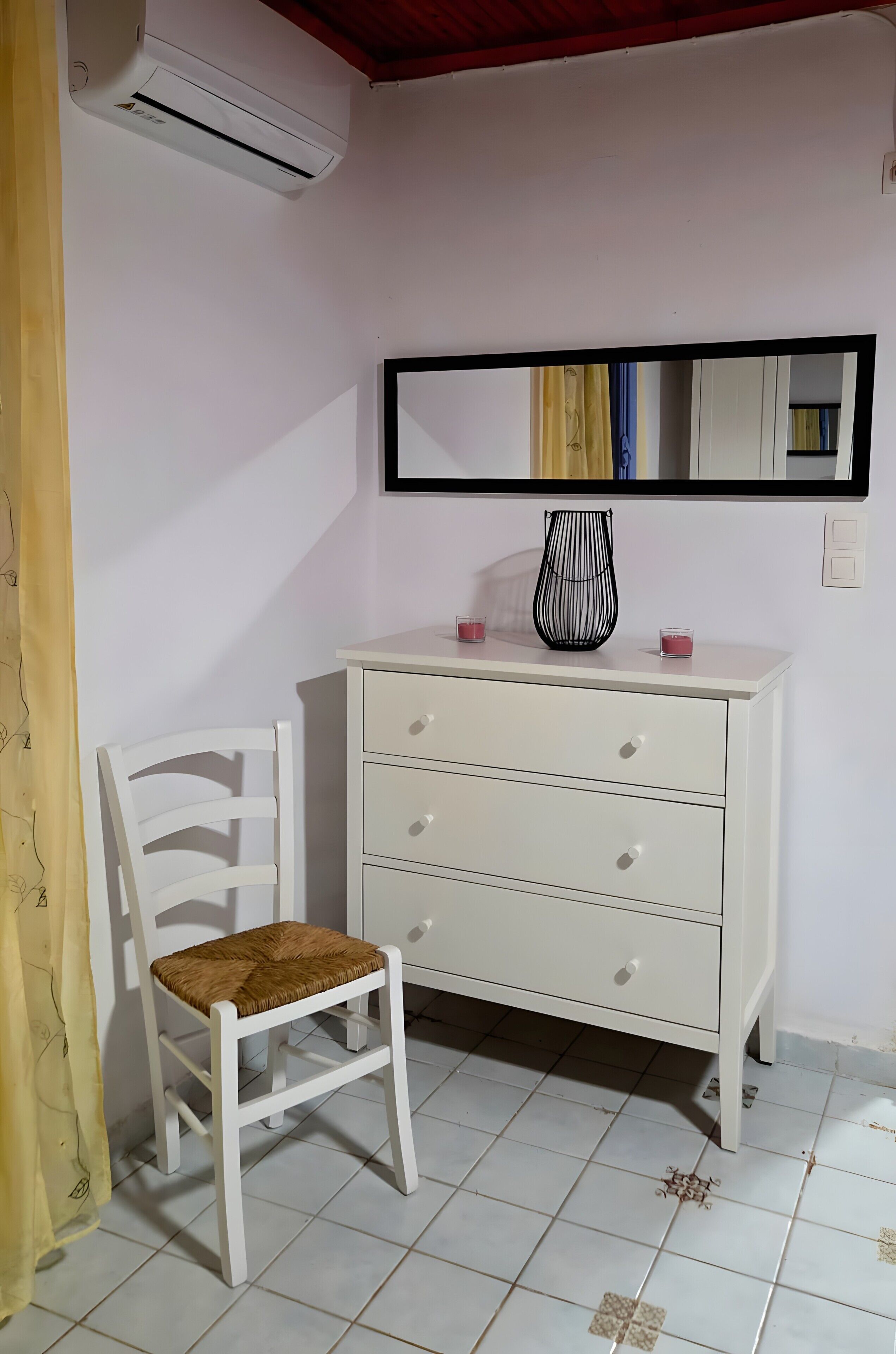 A white chair with a woven seat, a white dresser with drawers, a mirror above the dresser, and a tiled floor.