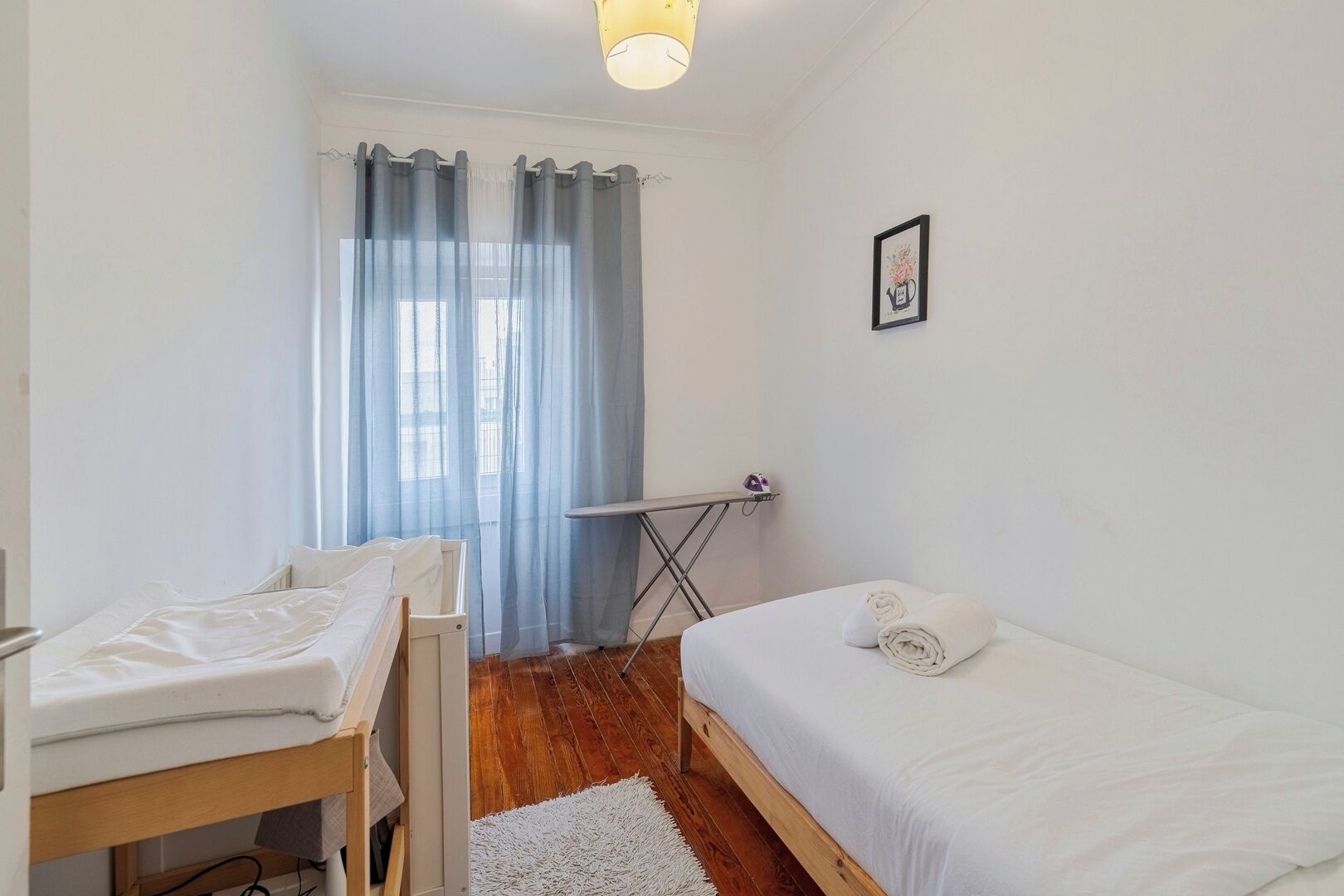A bedroom with a bed, a small table, a chair, a window with curtains, and a wall-mounted picture.