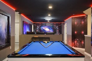 Ein Billardtisch mit blauer Spielbahn, umgeben von Wänden mit Weltraumkunst und roter Neonbeleuchtung.