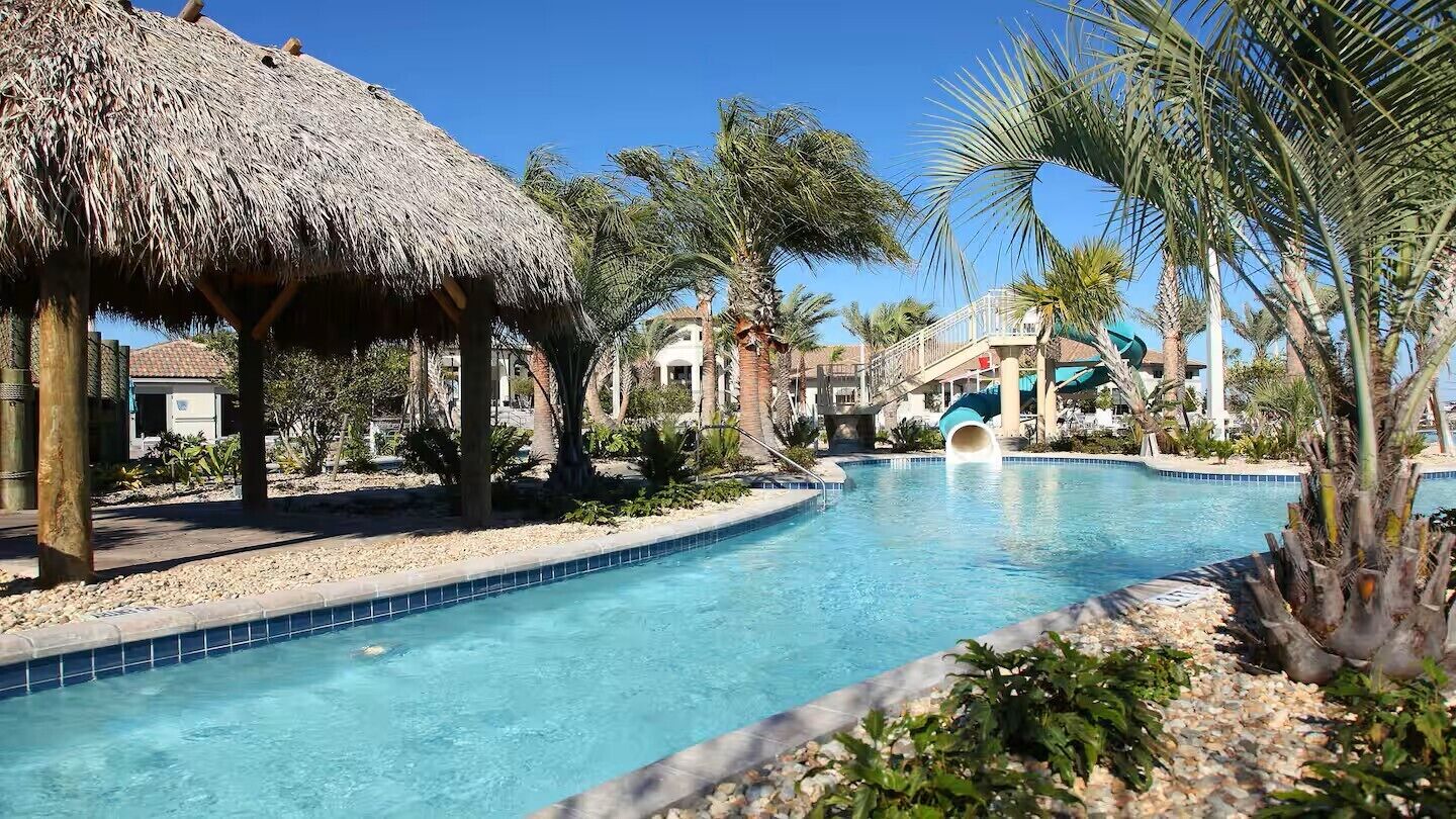 A swimming pool with a slide, surrounded by palm trees and a thatched-roof structure.