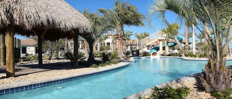 A swimming pool with a slide, surrounded by palm trees and a thatched-roof structure.