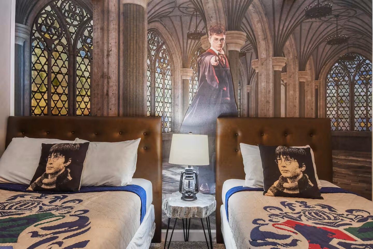 Une chambre avec deux lits, chacun orné d’un oreiller sur lequel est imprimé le visage d’un personnage. Une table avec une lampe se trouve entre les deux lits. La chambre présente un style architectural gothique, avec des fenêtres en arc et un plafond haut.
