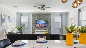 A modern dining area with a flat-screen TV mounted on a textured wall, a ceiling fan, and a table set for dining with blue placemats. - Storey Lake 8BR Family Adventure | Pool, Spa, Theater & Themed Rooms (Kissimmee)