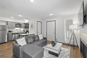 A modern living room with a grey sectional sofa, a coffee table, and a kitchen in the background. - Modern King Studio, Pool, Near MD Anderson and NRG (Houston)