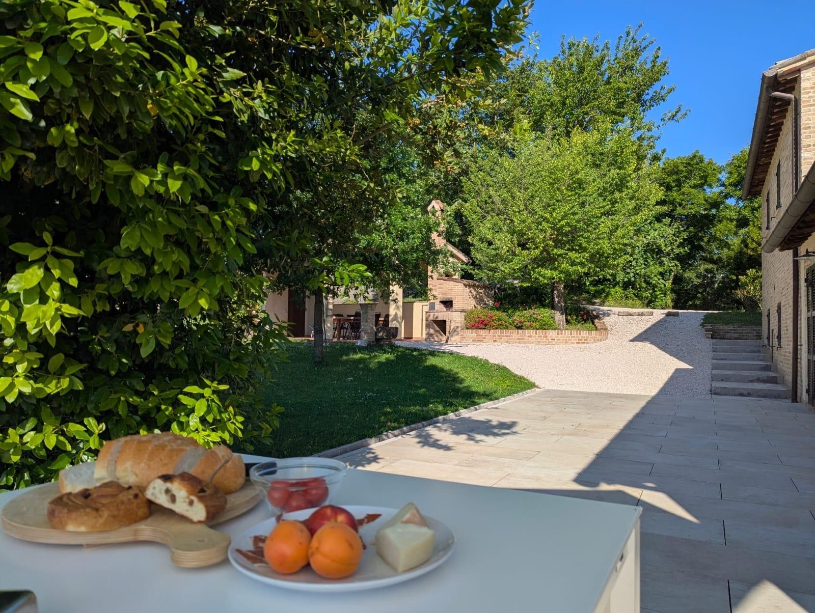 Un área de comedor al aire libre con una mesa dispuesta para una comida, rodeada de follaje y un edificio al fondo.