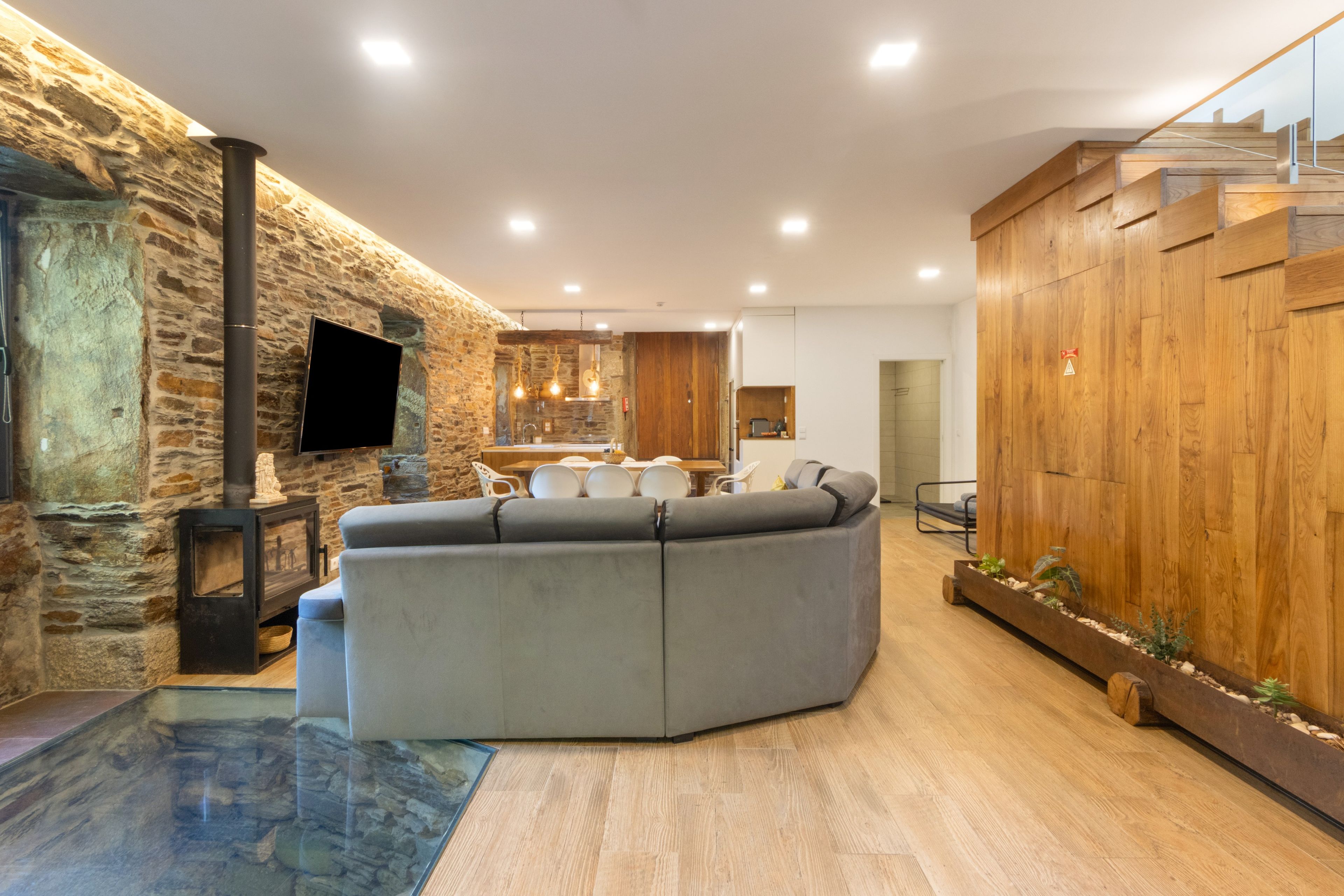 Un salon moderne avec un mur en pierre, un canapé d’angle, un téléviseur à écran plat et un escalier en bois.